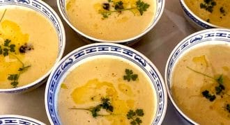 Several bowls of creamy yellow soup are garnished with fresh herbs and a drizzle of oil, served in blue and white patterned bowls, arranged closely together on a table.