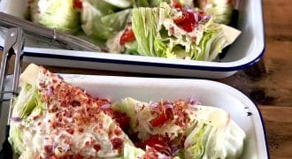 Trays of wedge salads topped with creamy dressing, diced tomatoes, bacon pieces, chopped red onion, and chives, served on white enamel trays with metal tongs on a wooden table.